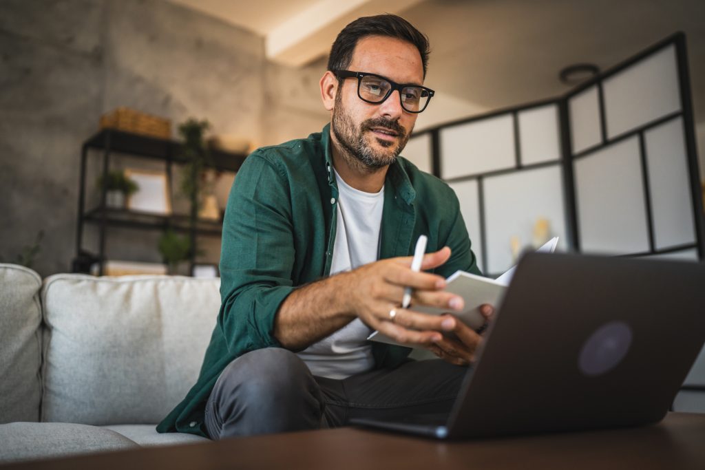 Adult man wear eyeglasses and a green shirt work from home in a modern living room, hold a notepad while use a laptop, suggesting remote work and creativity